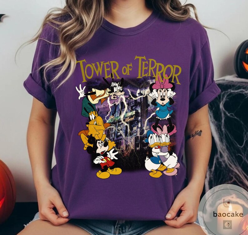 May include: Purple t-shirt featuring a graphic of Disney characters, including Mickey Mouse, Goofy, Minnie Mouse, and Donald Duck, with the text "Tower of Terror" in gold lettering. The design has a spooky, Halloween theme.