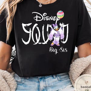 Puede incluir: Camiseta negra con el texto blanco "Disney Squad", con un dibujo de Daisy Duck con un kimono sosteniendo un globo de arco&iacute;ris. Las palabras "Big Sis" est&aacute;n debajo.