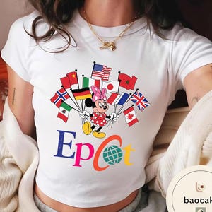 May include: White t-shirt with a Minnie Mouse design holding flags from different nations. The word "Epcot" is displayed in colourful letters, along with a globe graphic. Minnie Mouse is dressed in a red polka dot dress and a pink bow.