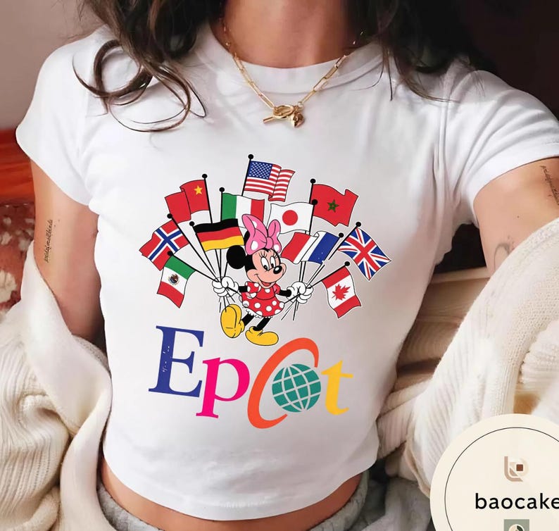 May include: White t-shirt with a Minnie Mouse design holding flags from different nations. The word "Epcot" is displayed in colourful letters, along with a globe graphic. Minnie Mouse is dressed in a red polka dot dress and a pink bow.