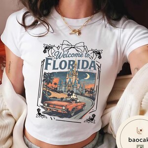 May include: White t-shirt featuring a graphic of Mickey Mouse driving a red convertible in front of a castle, with the text "Welcome to Florida EST. 1971". The design includes a bow and floral accents.