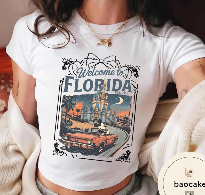 May include: White t-shirt featuring a graphic of Mickey Mouse driving a red convertible in front of a castle, with the text "Welcome to Florida EST. 1971". The design includes a bow and floral accents.