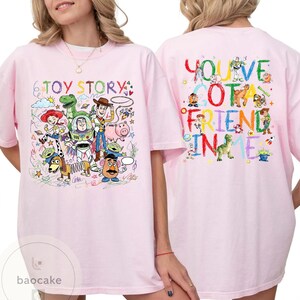 May include: Light pink t-shirt featuring colorful cartoon characters and the text "TOY STORY" on the front. The back of the shirt displays the phrase "YOU'VE GOT A FRIEND IN ME" with more cartoon characters.