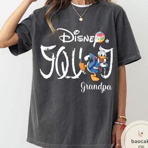 Puede incluir: Camiseta gris oscuro con la inscripci&oacute;n blanca "Disney Grandpa". La camiseta presenta un gr&aacute;fico colorido de Pato Donald con un atuendo azul. Una bola de arco&iacute;ris y una peque&ntilde;a flor tambi&eacute;n est&aacute;n incluidas en el dise&ntilde;o.