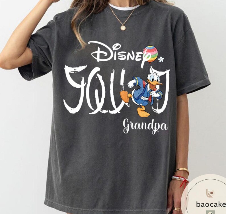 Puede incluir: Camiseta gris oscuro con la inscripci&oacute;n blanca "Disney Grandpa". La camiseta presenta un gr&aacute;fico colorido de Pato Donald con un atuendo azul. Una bola de arco&iacute;ris y una peque&ntilde;a flor tambi&eacute;n est&aacute;n incluidas en el dise&ntilde;o.