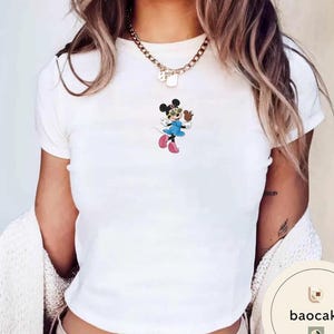 May include: A white short-sleeved t-shirt featuring a small graphic of a cartoon mouse wearing yellow sunglasses, a blue dress, and pink shoes. The mouse is holding a small brown object. A gold-toned chain necklace rests around the neckline.