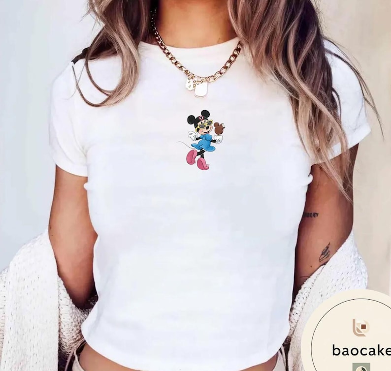 May include: A white short-sleeved t-shirt featuring a small graphic of a cartoon mouse wearing yellow sunglasses, a blue dress, and pink shoes. The mouse is holding a small brown object. A gold-toned chain necklace rests around the neckline.