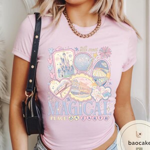 May include: Pale pink t-shirt with a colourful graphic design featuring a castle, a sphere, and other illustrations. The text "MAGICAL PLACE ON EARTH" is displayed in a whimsical font. The shirt has short sleeves and a round neckline.