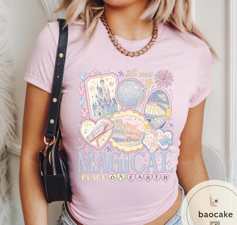 May include: Pale pink t-shirt with a colourful graphic design featuring a castle, a sphere, and other illustrations. The text "MAGICAL PLACE ON EARTH" is displayed in a whimsical font. The shirt has short sleeves and a round neckline.