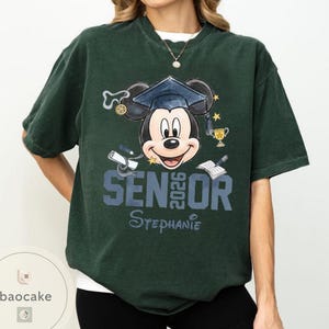 May include: Dark green t-shirt featuring a Mickey Mouse graphic wearing a graduation cap. The design includes the text "SENIOR 2026" and the name "Stephanie". Additional elements include a diploma, trophy, and open book.