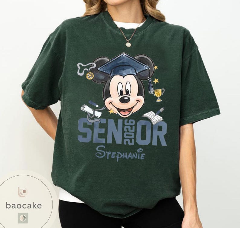 May include: Dark green t-shirt featuring a Mickey Mouse graphic wearing a graduation cap. The design includes the text "SENIOR 2026" and the name "Stephanie". Additional elements include a diploma, trophy, and open book.