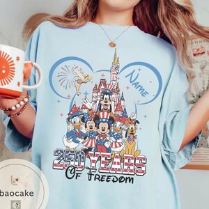 Peut inclure: T-shirt bleu clair avec un motif color&eacute; de personnages Disney devant un ch&acirc;teau, avec le texte "250 Years of Freedom". Le motif comprend Mickey Mouse, Donald Duck et d'autres personnages. Un nom peut &ecirc;tre ajout&eacute; au motif.