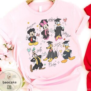 May include: Light pink t-shirt featuring cartoon characters Mickey Mouse, Minnie Mouse, Donald Duck, Daisy Duck, Goofy, and Pluto in graduation attire. Each character is wearing a graduation cap and gown. The shirt has character signatures.