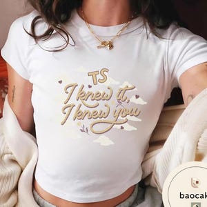 K&ouml;nnte beinhalten: Ein wei&szlig;es T-Shirt mit kurzen &Auml;rmeln und einem Grafikdesign mit dem Text "TS I knew it, I knew you" in einer Schriftart, die wie ein Seil aussieht. Das Design enth&auml;lt helle Wolkenillustrationen, kleine dunkle Herzen und ein kleines Blumenmotiv.