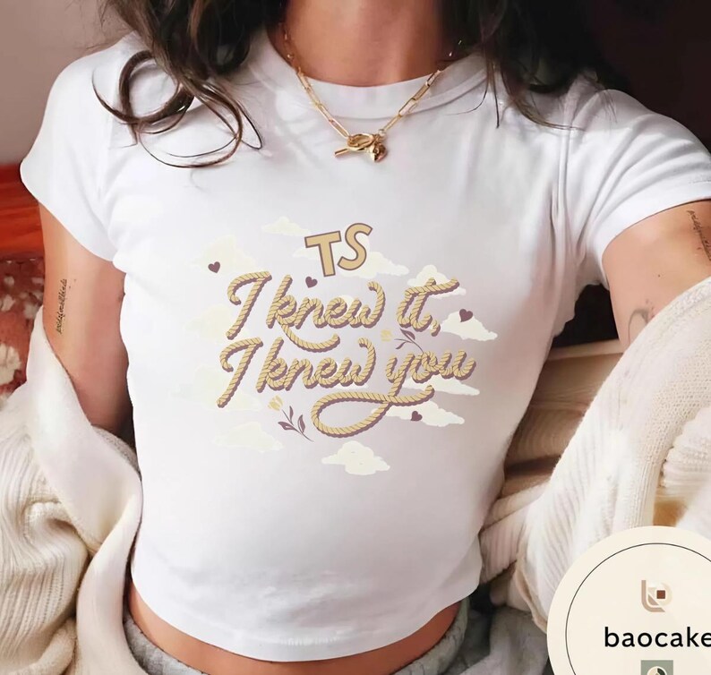 K&ouml;nnte beinhalten: Ein wei&szlig;es T-Shirt mit kurzen &Auml;rmeln und einem Grafikdesign mit dem Text "TS I knew it, I knew you" in einer Schriftart, die wie ein Seil aussieht. Das Design enth&auml;lt helle Wolkenillustrationen, kleine dunkle Herzen und ein kleines Blumenmotiv.