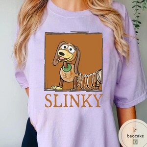 K&ouml;nnte beinhalten: Ein hellviolettes T-Shirt mit einer rechteckigen Grafik eines braunen Zeichentrickhundes mit federartigem K&ouml;rper und gr&uuml;nem Halsband. Der Hund ist vor einem braunen Hintergrund abgebildet, darunter steht das Wort SLINKY in gro&szlig;en Buchstaben.
