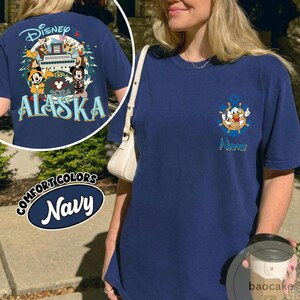 May include: Navy blue t-shirt with a Disney Alaska 2026 graphic on the back, featuring Mickey Mouse, Donald Duck, and other characters. The front of the shirt has a Donald Duck anchor design. The shirt is labeled "Comfort Colors Navy".