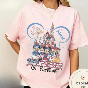 Peut inclure: T-shirt rose clair avec un motif color&eacute; de personnages Disney en tenues patriotiques. Le design comprend un ch&acirc;teau, des feux d'artifice et le texte "250 Years of Freedom" et "Name". Le t-shirt a des manches courtes et un col rond.
