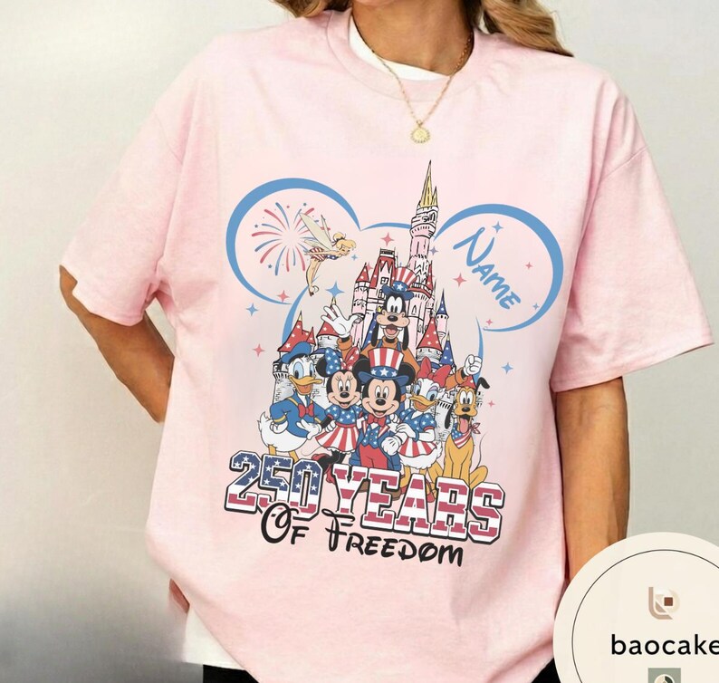 Peut inclure: T-shirt rose clair avec un motif color&eacute; de personnages Disney en tenues patriotiques. Le design comprend un ch&acirc;teau, des feux d'artifice et le texte "250 Years of Freedom" et "Name". Le t-shirt a des manches courtes et un col rond.
