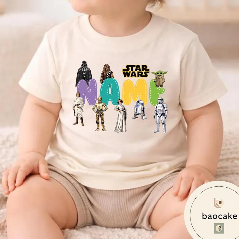 May include: Cream-colored Star Wars themed t-shirt featuring the word "NAME" in colorful letters with Star Wars characters. The shirt has a crew neck and short sleeves. The shirt is for babies and toddlers.