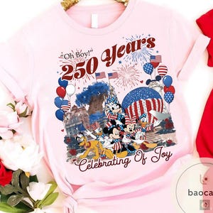 Puede incluir: Una camiseta rosa claro con un gr&aacute;fico colorido que celebra 250 a&ntilde;os. El dise&ntilde;o incluye Mickey Mouse, Minnie Mouse, Donald Duck y Pluto, junto con fuegos artificiales, globos y banderas americanas. El texto incluye "Oh Boy!" y "Celebrating Of Joy."