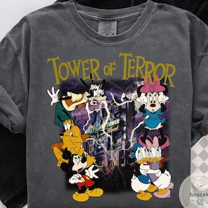 May include: Dark gray t-shirt featuring the text "Tower of Terror" in gold. The graphic includes Mickey Mouse, Goofy, Pluto, Minnie Mouse, and Donald Duck in front of a haunted hotel with lightning.