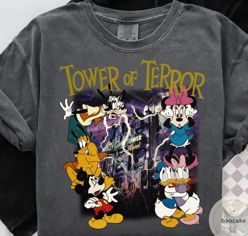 May include: Dark gray t-shirt featuring the text "Tower of Terror" in gold. The graphic includes Mickey Mouse, Goofy, Pluto, Minnie Mouse, and Donald Duck in front of a haunted hotel with lightning.