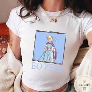 K&ouml;nnte beinhalten: Ein wei&szlig;es T-Shirt mit kurzen &Auml;rmeln und einer grafischen Illustration von Bo Peep in einem blauen quadratischen Rahmen. Die Figur tr&auml;gt einen blauen Overall, eine rosa Sch&auml;rpe und einen violetten Umhang und h&auml;lt einen Hirtenstab. Der Text BO PEEP steht darunter.