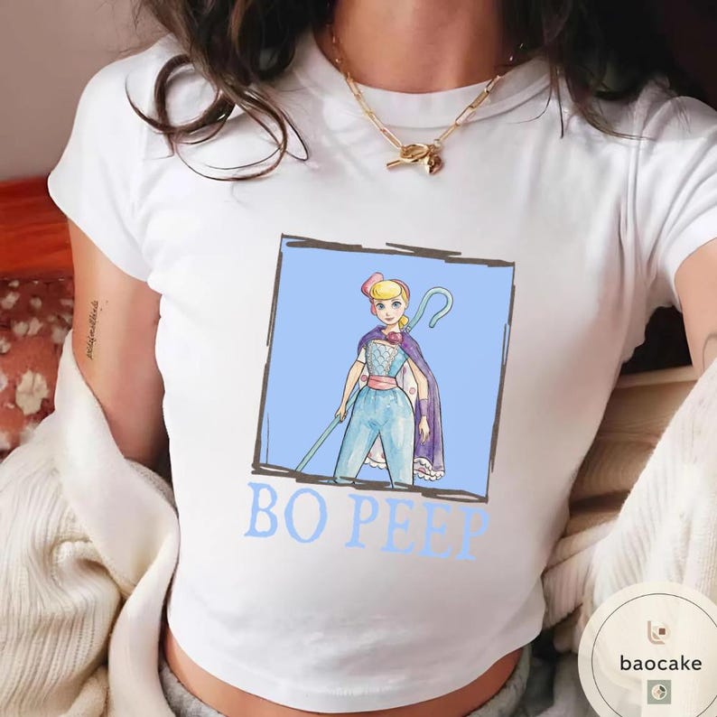 K&ouml;nnte beinhalten: Ein wei&szlig;es T-Shirt mit kurzen &Auml;rmeln und einer grafischen Illustration von Bo Peep in einem blauen quadratischen Rahmen. Die Figur tr&auml;gt einen blauen Overall, eine rosa Sch&auml;rpe und einen violetten Umhang und h&auml;lt einen Hirtenstab. Der Text BO PEEP steht darunter.