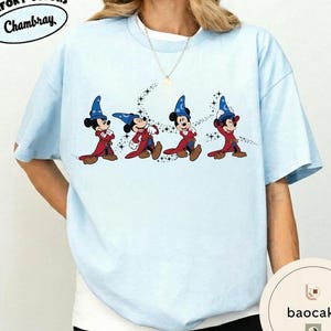 May include: Light blue t-shirt featuring a graphic of Mickey Mouse in wizard attire. The design shows Mickey in a red robe and blue wizard hat, with a trail of stars. The shirt has the text "Chambray" in a circular design.