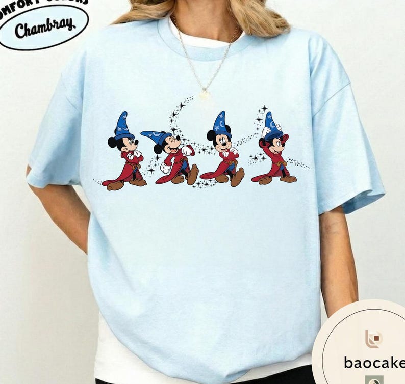 May include: Light blue t-shirt featuring a graphic of Mickey Mouse in wizard attire. The design shows Mickey in a red robe and blue wizard hat, with a trail of stars. The shirt has the text "Chambray" in a circular design.