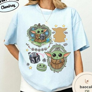 May include: A light blue t-shirt featuring a graphic print of a green creature with large eyes, a silver helmet, a green macaron, and the text GROGU. The design includes decorative frames, small stars, and blue butterflies on a light blue fabric.