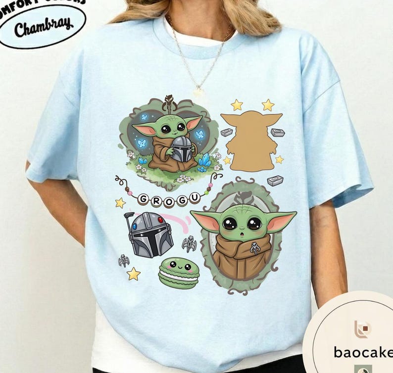 May include: A light blue t-shirt featuring a graphic print of a green creature with large eyes, a silver helmet, a green macaron, and the text GROGU. The design includes decorative frames, small stars, and blue butterflies on a light blue fabric.