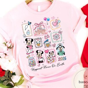 May include: A light pink t-shirt featuring a collage of Disney characters in nurse attire. The design includes Mickey Mouse, Minnie Mouse, Donald Duck, Daisy Duck, and Goofy, along with the text "Happiest Nurse On Earth" and the year "2026".
