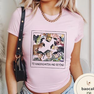 May include: A pale pink t-shirt with a framed graphic of animated characters wearing graduation caps. The text "TO KINDERGARTEN AND BEYOND" is printed below the graphic. The shirt has short sleeves.