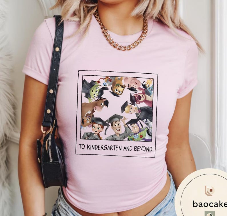 May include: A pale pink t-shirt with a framed graphic of animated characters wearing graduation caps. The text "TO KINDERGARTEN AND BEYOND" is printed below the graphic. The shirt has short sleeves.