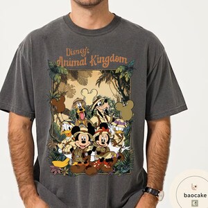 May include: Dark grey t-shirt featuring a graphic of Disney's Animal Kingdom characters, including Mickey and Minnie Mouse, Donald Duck, and Goofy, in safari attire. The shirt has the text "Disney's Animal Kingdom" in a jungle-themed design.