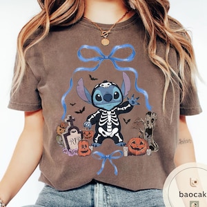 May include: Brown t-shirt featuring a Halloween-themed Stitch design. The graphic includes Stitch in a skeleton costume, pumpkins, bats, and a tombstone. Blue bows and ribbons frame the central image.