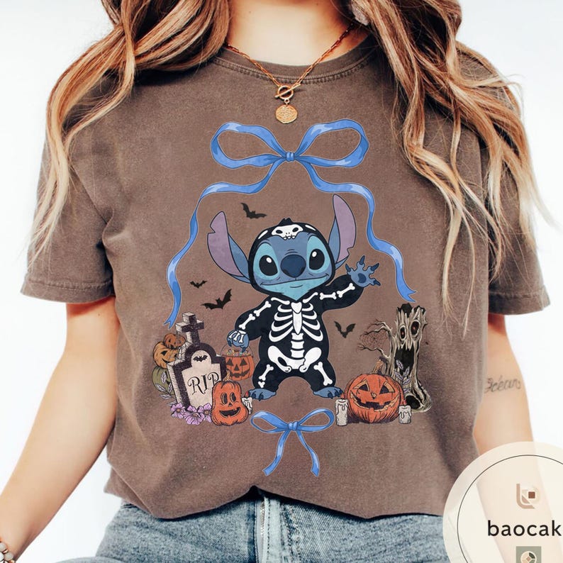 May include: Brown t-shirt featuring a Halloween-themed Stitch design. The graphic includes Stitch in a skeleton costume, pumpkins, bats, and a tombstone. Blue bows and ribbons frame the central image.