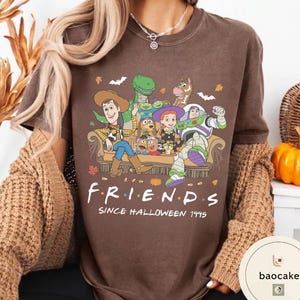 May include: A brown short-sleeve t-shirt featuring a graphic of Toy Story characters sitting on a couch with the text "FRIENDS SINCE HALLOWEEN 1995". The design includes Woody, Buzz Lightyear, Jessie, Rex, Slinky Dog, and Mr. Potato Head.