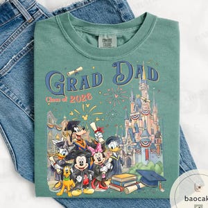 May include: Green Comfort Colors t-shirt with a graphic featuring Disney characters, the text "Grad Dad Class of 2026", and a castle illustration. The shirt is folded on a pair of blue jeans.