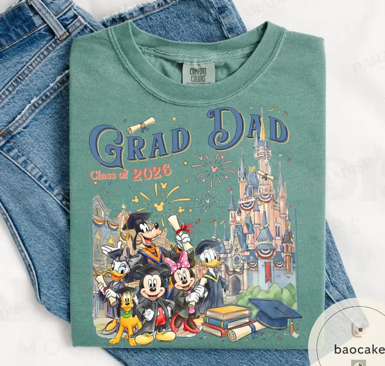 May include: Green Comfort Colors t-shirt with a graphic featuring Disney characters, the text "Grad Dad Class of 2026", and a castle illustration. The shirt is folded on a pair of blue jeans.