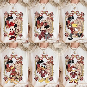 May include: Cream-colored t-shirts featuring various Disney characters in a framed design. Each design includes a character like Minnie Mouse, Mickey Mouse, Goofy, Pluto, or Donald Duck, with a building and floral accents. The designs are in shades of red, gold, and black.