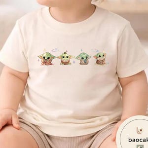 May include: Cream-colored t-shirt featuring a graphic of four cartoon characters. The characters are green with large eyes and are in different poses. The shirt is made of a soft material and is suitable for children.