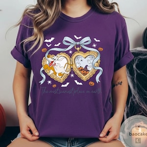 May include: A purple t-shirt with a Halloween design. The design features a heart-shaped locket with cartoon characters, pumpkins, bats, and spiders. The text "The most scariest place on earth" is printed below the locket.