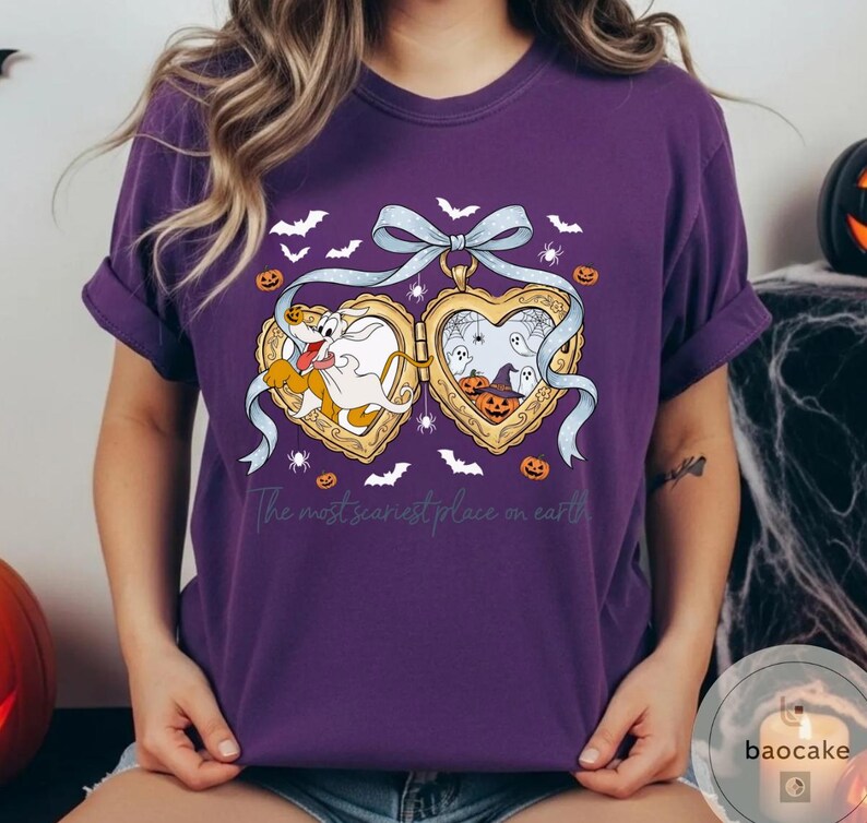May include: A purple t-shirt with a Halloween design. The design features a heart-shaped locket with cartoon characters, pumpkins, bats, and spiders. The text "The most scariest place on earth" is printed below the locket.