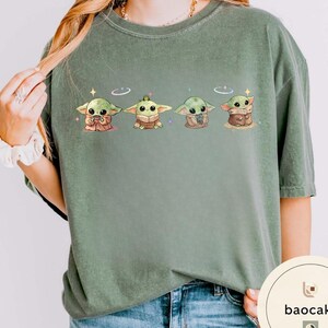 May include: Sage green t-shirt featuring four cartoon illustrations of a Grogu character. Each Grogu is in a different pose, with a sparkling star above each head. The shirt is made of a soft material.