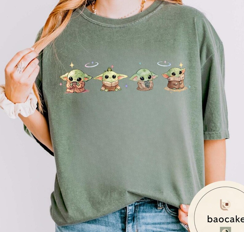 May include: Sage green t-shirt featuring four cartoon illustrations of a Grogu character. Each Grogu is in a different pose, with a sparkling star above each head. The shirt is made of a soft material.