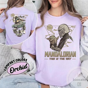 Op de afbeelding: Een lichtpaars T-shirt met een afbeelding van The Mandalorian en Grogu. Het ontwerp bevat de tekst "MANDALORIAN" en "THIS IS THE WAY". De achterkant van het shirt heeft ook een afbeelding met de tekst "MANDALORIAN GROGU".