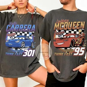 Puede incluir: Dos camisetas gris carb&oacute;n con gr&aacute;ficos de la pel&iacute;cula "Cars". Una camiseta muestra un coche azul con el texto "Sally Carrera" y "Think Fast 301". La otra camiseta muestra un coche rojo con "Lightning McQueen" y "Think Fast 95."